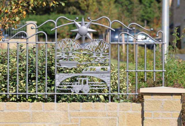 The panel has a large sun above the Forth Bridge, with a boat sailing on the watery Firth of Forth below, then a modern train crossing a bridge and finally a tractor, representing the architecture of Inverkeithing High School, in a ploughed field.