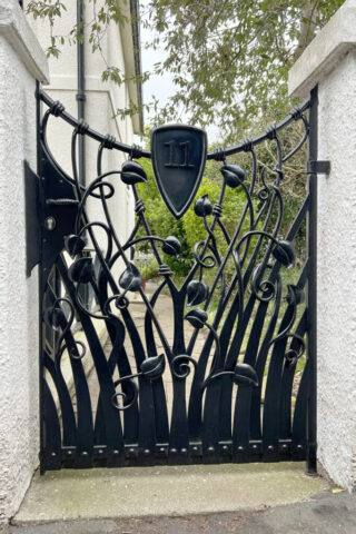 Black gloss forged gate set in a white wall. The gate is made up of forged elements of leaves and tendrils interwoven to give a pleasing design.