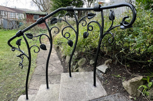 Handrail to three steps in a back garden. The handrail is forged from mild steel with forged leaves and tapered tendrils coming from the posts. The posts are attached to the handrail by wrapping the steel around. The metalwork is painted gloss black.