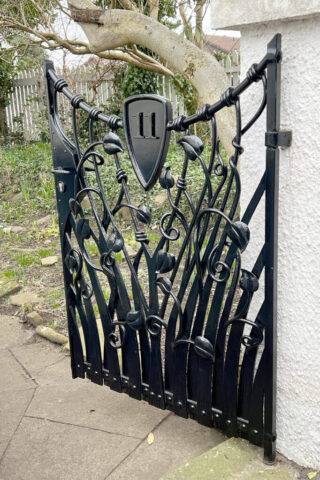 Black gloss forged gate set in a white wall. The gate is made up of forged elements of leaves and tendrils interwoven to give a pleasing design.