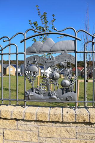 Forged and fabricated panel made from mild steel, painted a soft grey colour. The panel shows mountains dropping to an under the sea view of fish, sea weed, a jelly fish and rocks.