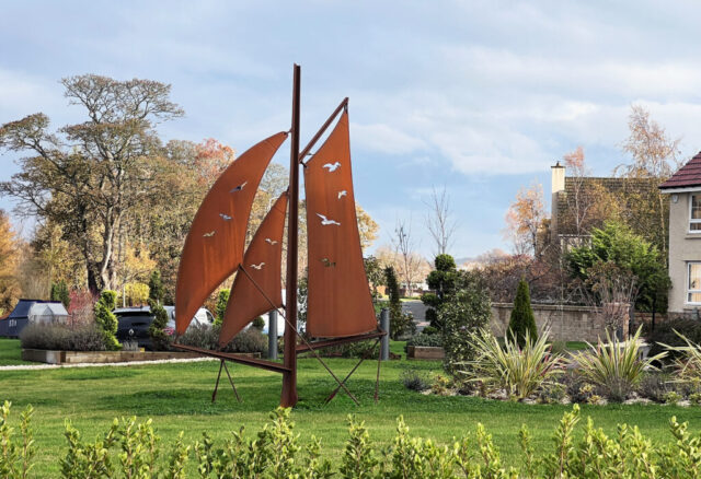Large sculpture with three sails with gulls pierced through the sails. The sculpture is created from CorTen steel which weathers to a rusty brown finish. The sculpture sits within a grassed landscaped area.