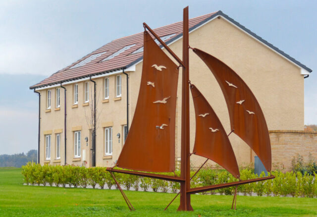 Large sculpture with three sails with gulls pierced through the sails. The sculpture is created from CorTen steel which weathers to a rusty brown finish. The sculpture sits within a grassed landscaped area.