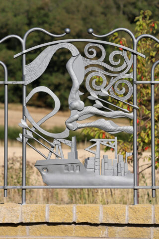 A decorative panel forged and fabricated from mild steel, depicting a stylised face, a cargo ship, arrows and swirling elements representing the blowing wind.