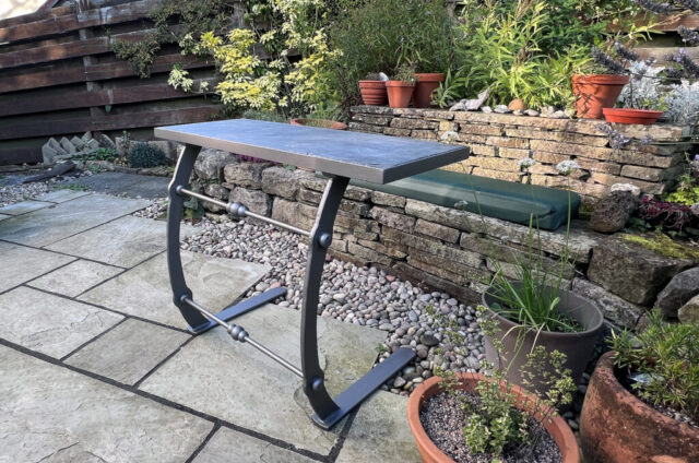 Two legged forged table sitting on slabs on a garden patio. The table has a slate top. The table is painted grey.