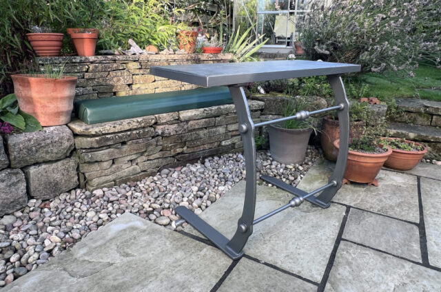 Two legged forged table sitting on slabs on a garden patio. The table has a slate top. The table is painted grey.