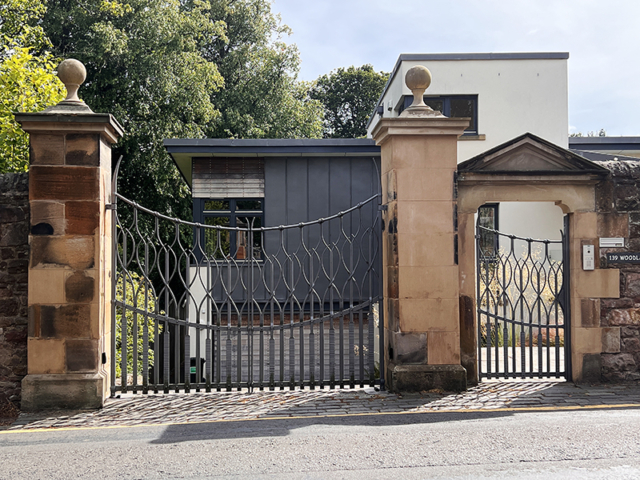 Double Gate and pedestrian gate attached to traditional old stone pillars leading to a modern home. The gate is forged from mild steel with heavy texturing on flat plates along the bottom half of the gates with round bars rising up to the top rail as they cross over forming a delicate pattern.