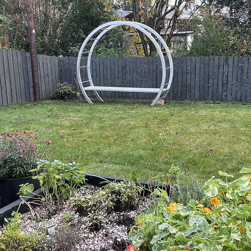 garden bench created with two large hoops that disappear into the ground so that the bench looks like a large moon rising. Slatted bench across to sit on. Painted silver.